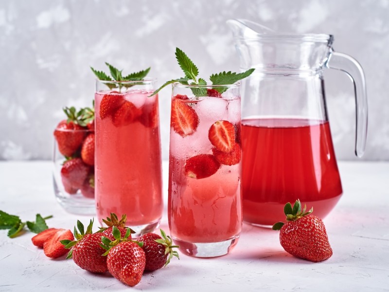 2 vasos de limonada de fresa con mango y plátano, junto a una jarra con la limonada y algunas fresas frescas.