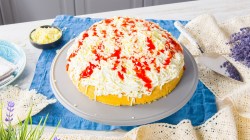Un pastel helado de espagueti en bandeja está sobre una mesa de madera. A su izquierda hay algunas fresas. A su derecha, una rebanadora de pastel.