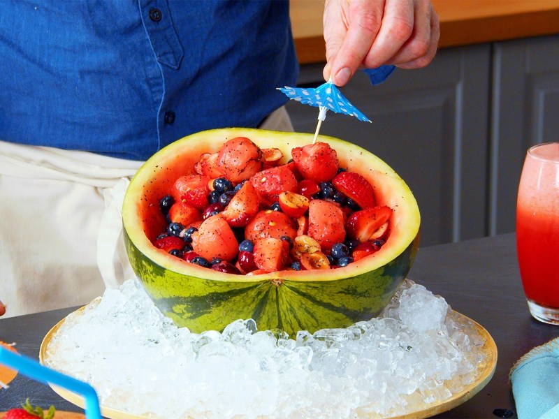 Una ensalada de frutas dentro de la cáscara de una sandía. La cáscara está colocada en un tazón grande con cubitos de hielo para que la ensalada se mantenga fresca. Se puede ver al chef Oli colocando una sombrillita de cóctel en la ensalada. A la derecha del tazón hay un vaso con un cóctel de sandía.