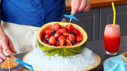 Una ensalada de frutas dentro de la cáscara de una sandía. La cáscara está colocada en un tazón grande con cubitos de hielo para que la ensalada se mantenga fresca. Se puede ver al chef Oli colocando una sombrillita de cóctel en la ensalada. A la derecha del tazón hay un vaso con un cóctel de sandía.