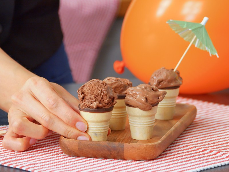 Una pequeña tabla de madera que está sobre un mantel individual a rayas rojas. En la tabla hay algunos barquillos con helado de chocolate casero. En una bola de helado hay una pequeña sombrilla de cóctel. Una mano agarra uno de los barquillos de helado. Al fondo se puede ver un globo naranja.
