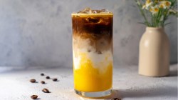 Un vaso grande con latte helado de mango, colocado sobre un fondo minimalista claro. Hay algunos granos de café esparcidos. Al fondo hay un jarrón beige con manzanilla.