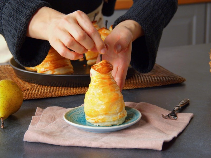 Una pera rellena envuelta en hojaldre, colocada en un plato pequeño por la cocinera Adrianna. En la imagen también se ve una pera fresca en la encimera, otra pera rellena. En el borde derecho de la imagen hay una cesta.
