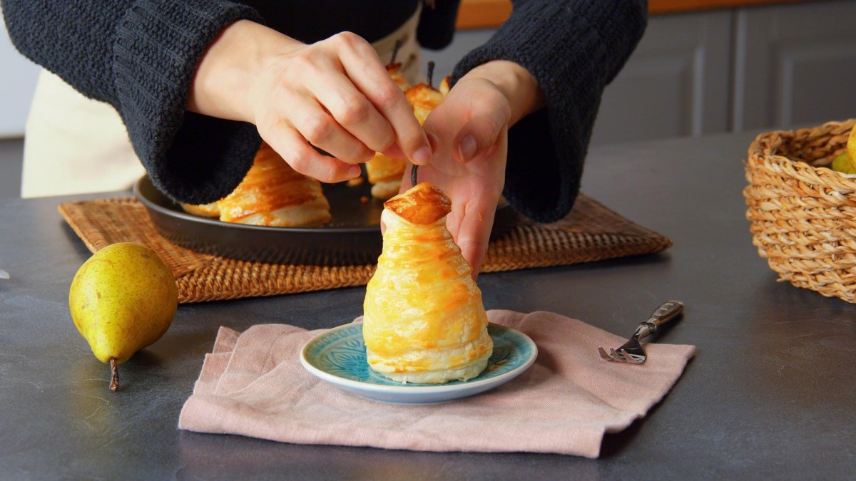 Una pera rellena envuelta en hojaldre, colocada en un plato pequeño por la cocinera Adrianna. En la imagen también se ve una pera fresca en la encimera, otra pera rellena. En el borde derecho de la imagen hay una cesta.