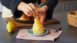 Una pera rellena envuelta en hojaldre, colocada en un plato pequeño por la cocinera Adrianna. En la imagen también se ve una pera fresca en la encimera, otra pera rellena. En el borde derecho de la imagen hay una cesta.
