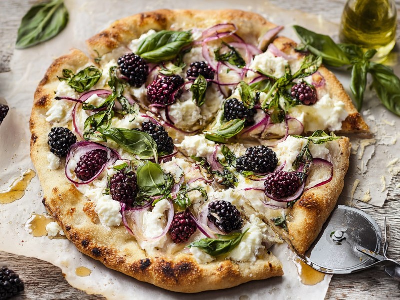 Una pizza de moras y ricota, adornada con albahaca fresca y cubierta adicionalmente con cebolla morada. La pizza está sobre papel para hornear y rociada con miel. Junto a ella hay un tazón con moras y otro con queso rallado. También se puede ver un cortador de pizza.