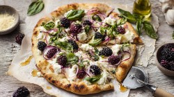 Una pizza de moras y ricota, adornada con albahaca fresca y cubierta adicionalmente con cebolla morada. La pizza está sobre papel para hornear y rociada con miel. Junto a ella hay un tazón con moras y otro con queso rallado. También se puede ver un cortador de pizza.