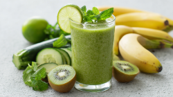 Un vaso de smoothie de kiwi y calabacín sobre una encimera gris. Alrededor del vaso hay kiwis, pepinos, plátanos y un calabacín. El smoothie está decorado con una rodaja de lima.